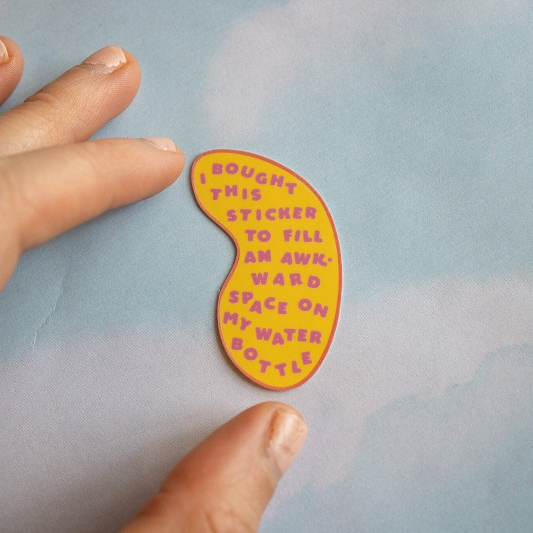 Hand holding a colorful sticker with text that says "I bought this sticker to fill an awkward space on my water bottle" on a light blue background