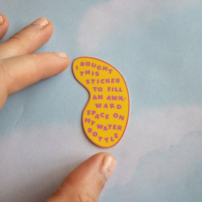 Hand holding a colorful sticker with text that says "I bought this sticker to fill an awkward space on my water bottle" on a light blue background