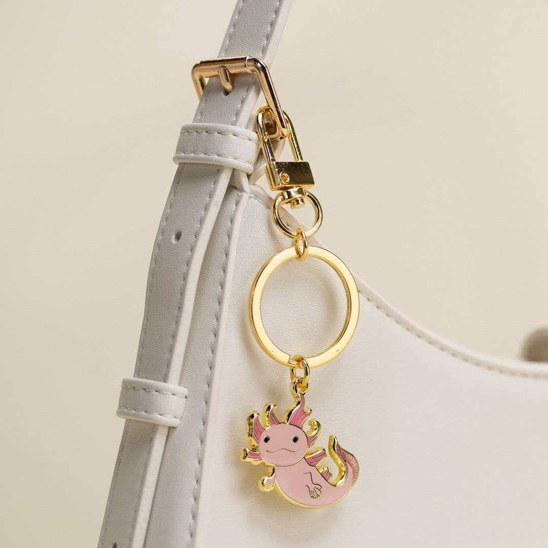 Pink axolotl keychain on a gold ring attached to a light gray strap.