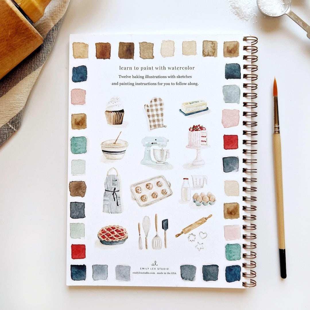 Spiral-bound art book with baking illustrations on a white surface with a paintbrush 