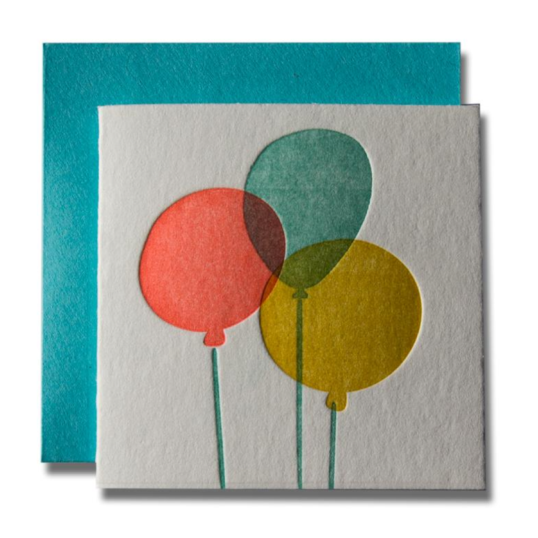 A square greeting card with a textured off-white surface featuring three overlapping watercolor-style balloons in coral red, teal, and mustard yellow, shown with a teal envelope behind it on a white background.