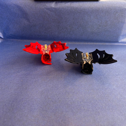 Two decorative bat shaped claw hair clips, one red and one glossy black, displayed on a textured blue background.