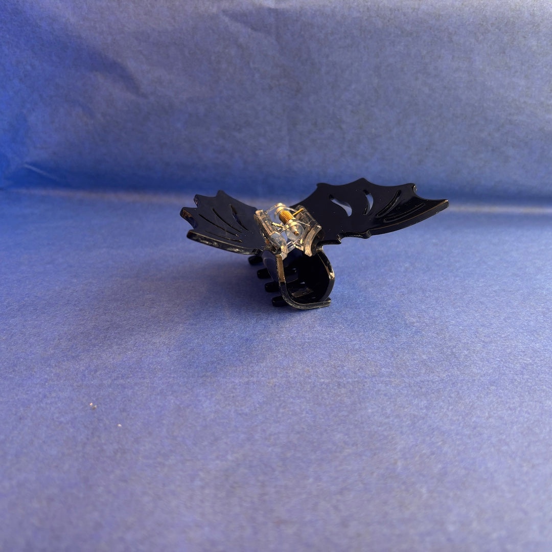 A black plastic bat shaped claw-style hair clip with a small gold-toned metal accent in the center, photographed against a blue textured background.