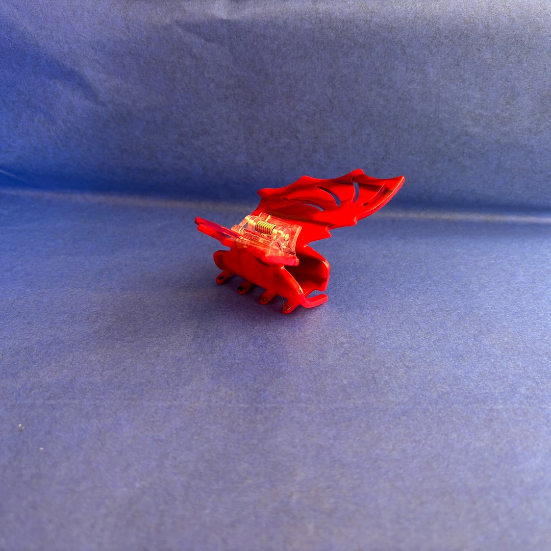 A small red plastic bat shaped claw clip with a clear, transparent center section, photographed on a textured blue background.
