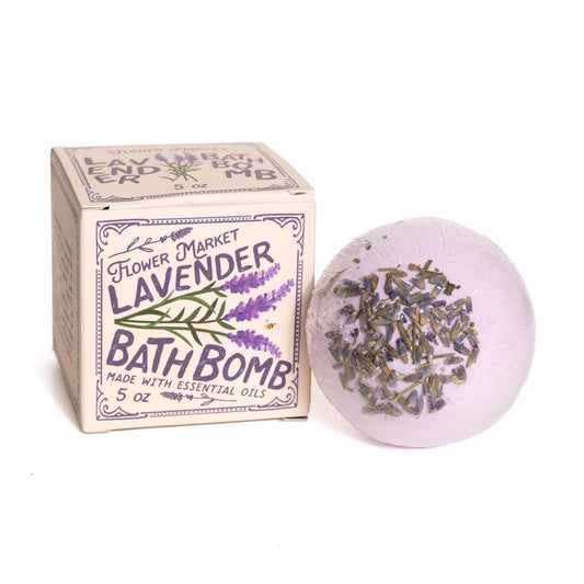A lavender bath bomb with dried lavender buds embedded inside, shown next to its illustrated paper box labeled 'Flower Market Lavender Bath Bomb' on a plain white background.