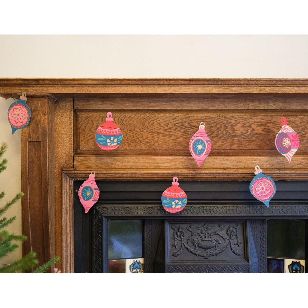 A set of colorful paper Christmas ornament garlands in pink, blue, and red hanging on a rustic wooden fireplace mantel.