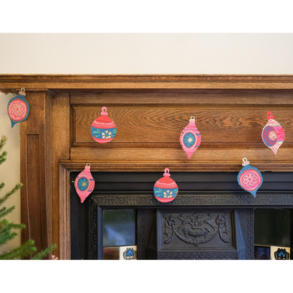A set of colorful paper Christmas ornament garlands in pink, blue, and red hanging on a rustic wooden fireplace mantel.