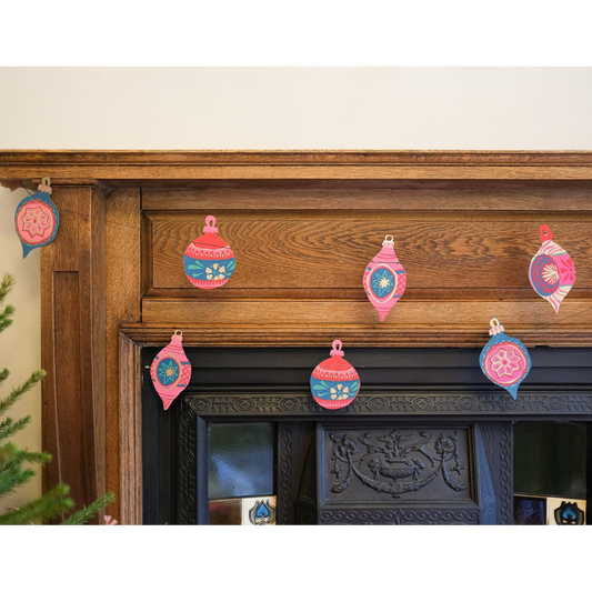 A set of colorful paper Christmas ornament garlands in pink, blue, and red hanging on a rustic wooden fireplace mantel.
