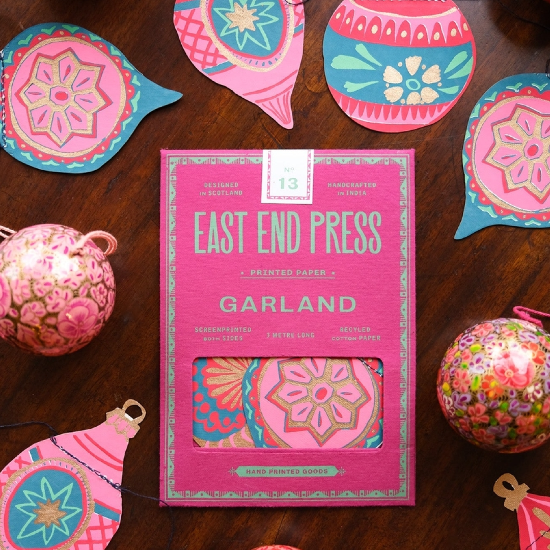 A pink East End Press Garland printed paper sheet with a window showing colorful patterned paper inside, styled on a dark wood surface with decorative paper ornaments around it.