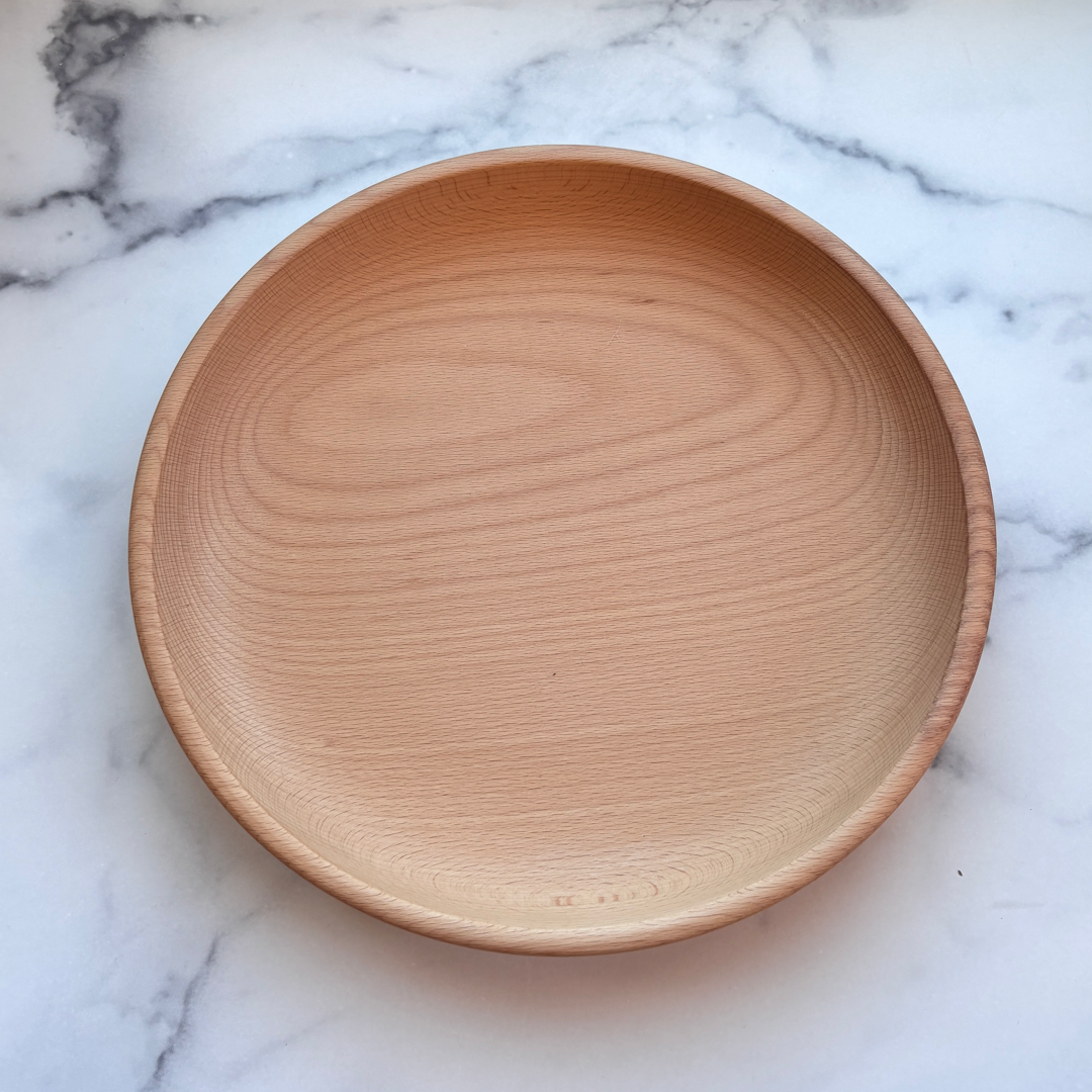 A round, shallow wooden serving bowl with a natural light oak finish, photographed from directly above on a white marble surface with gray veining.