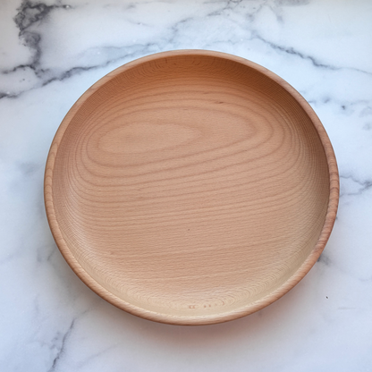 A round, shallow wooden serving bowl with a natural light oak finish, photographed from directly above on a white marble surface with gray veining.