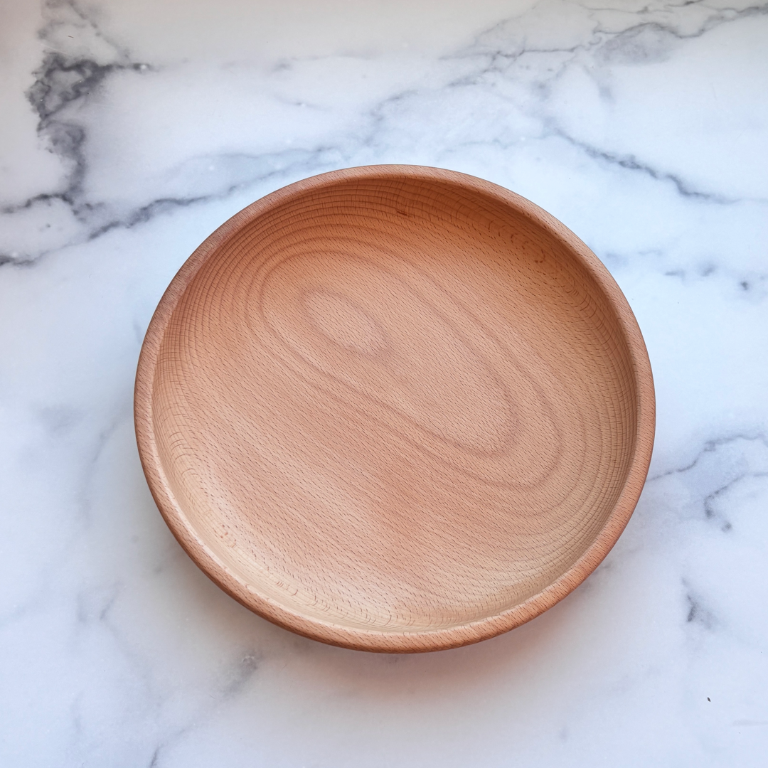 A round, shallow wooden plate with a natural light brown wood grain finish, photographed from directly above on a white marble surface with gray veining.