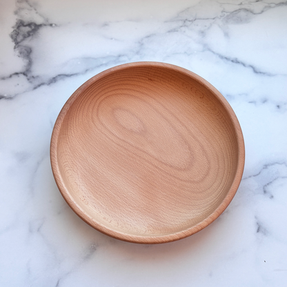 A round, shallow wooden plate with a natural light brown wood grain finish, photographed from directly above on a white marble surface with gray veining.