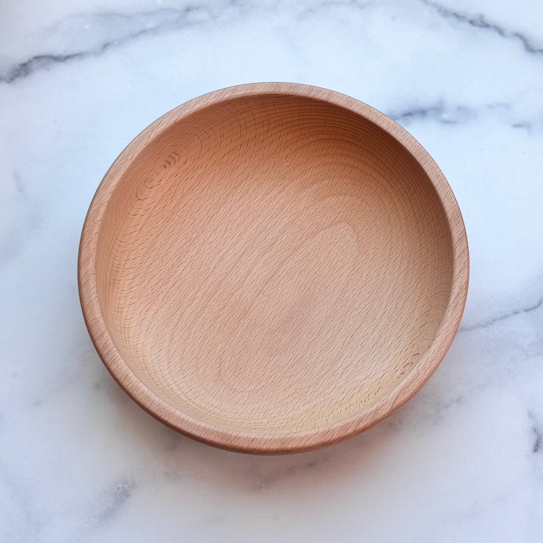A round, handcrafted wooden bowl with a natural light oak finish, photographed from directly above on a white and gray marble surface.