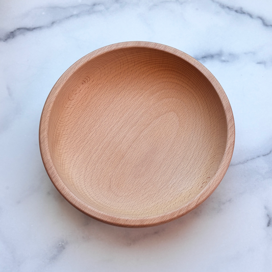 A round, handcrafted wooden bowl with a natural light oak finish, photographed from directly above on a white and gray marble surface.