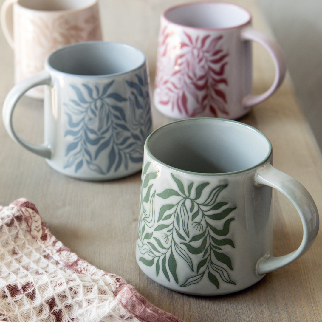 Three ceramic mugs with botanical leaf patterns in blue, green, and pink, arranged on a light wooden table with a textured knit cloth in the foreground.