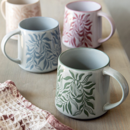 Three ceramic mugs with botanical leaf patterns in blue, green, and pink, arranged on a light wooden table with a textured knit cloth in the foreground.