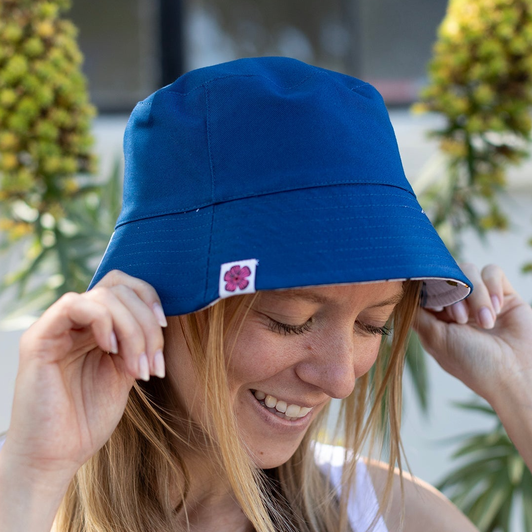 Woman wearing a blue bucket hat with a pink flower logo, standing outdoors.