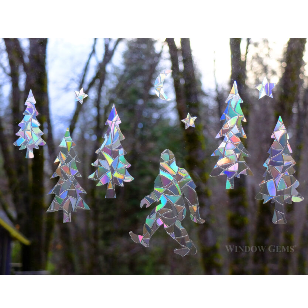 Iridescent suncatcher window decor featuring trees, bigfoot, a crescent moon, and stars shown hanging against a blurred forest background.