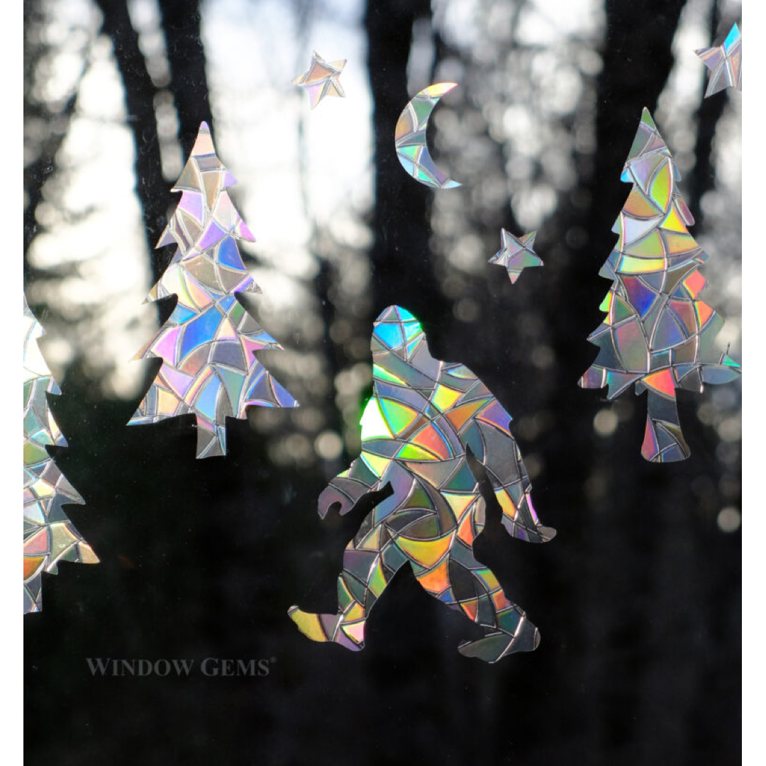 Iridescent suncatcher window decor featuring trees, bigfoot, a crescent moon, and stars shown hanging against a blurred forest background.