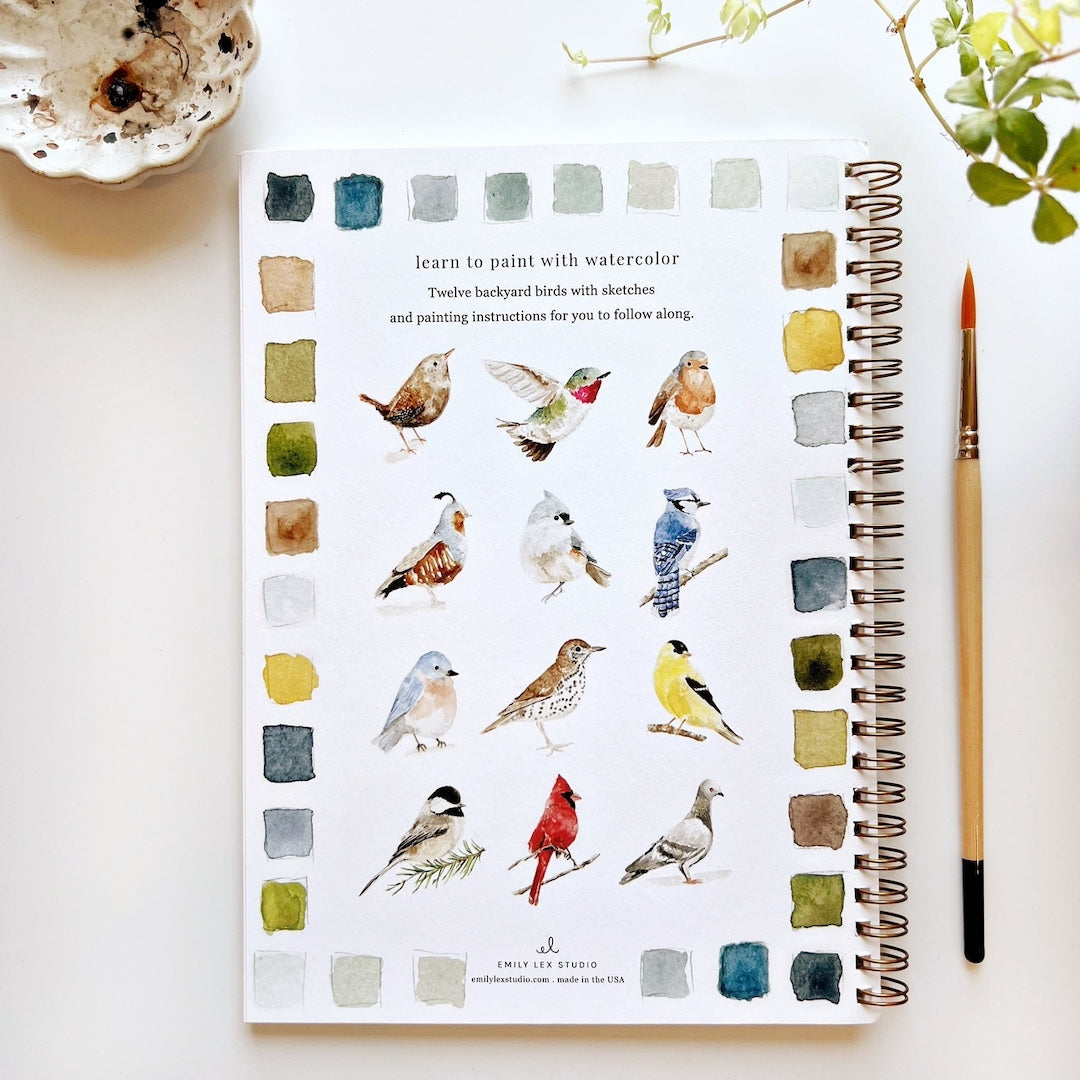 Artistic notebook with watercolor illustrations of birds and painting instructions on a white surface.