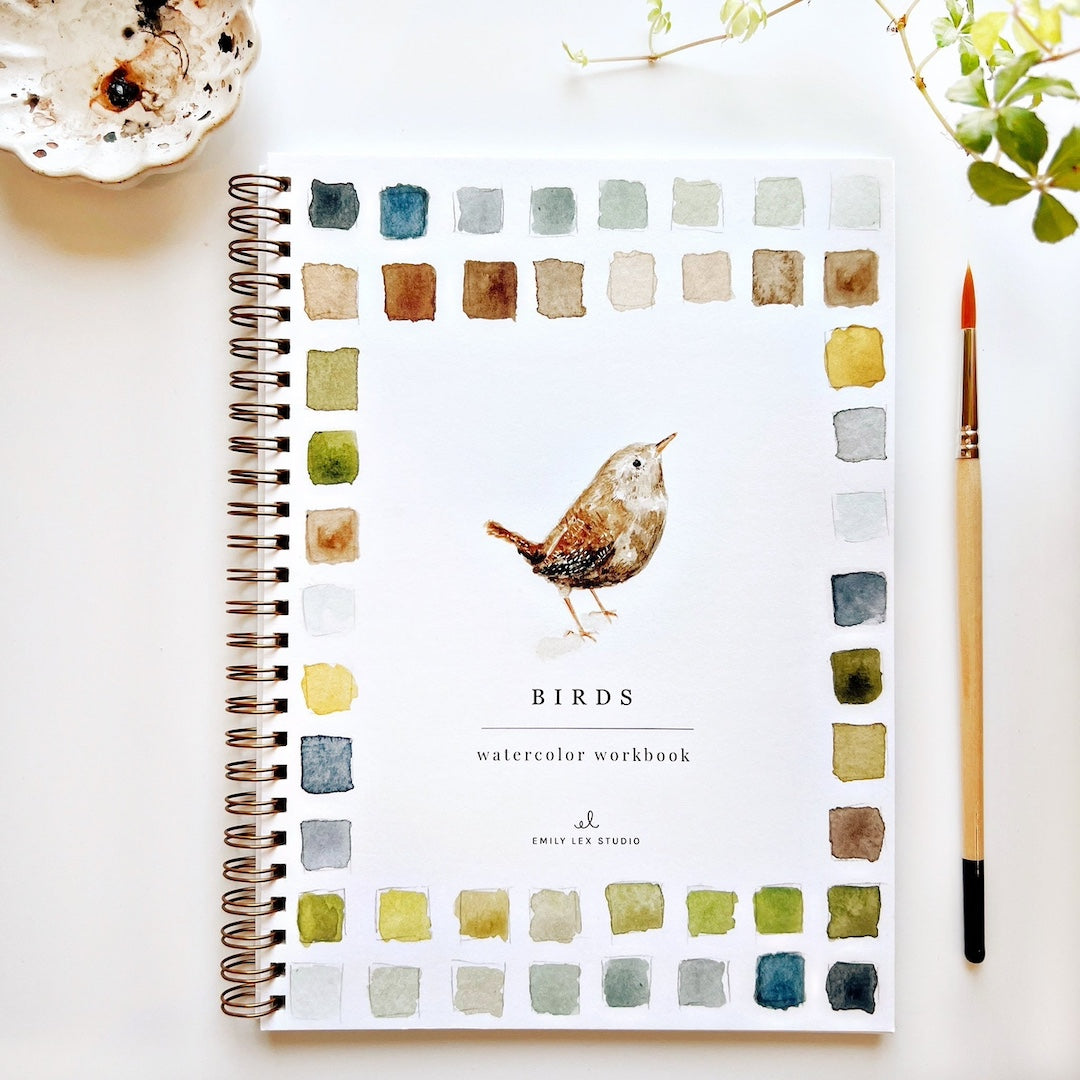 Watercolor workbook with bird illustration on a white surface with a cup and plant.