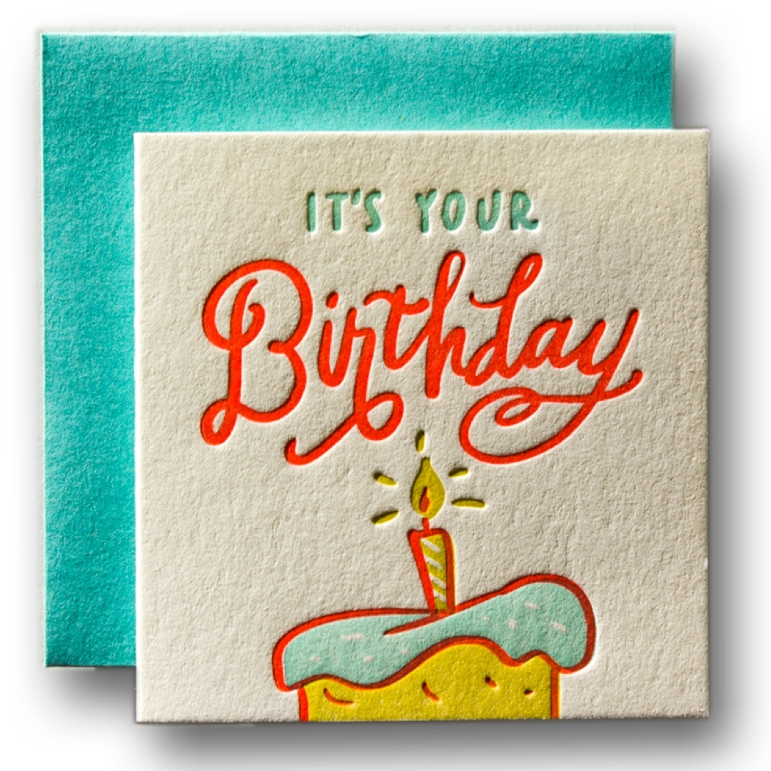 A square birthday card with a textured cream background, featuring the words 'IT'S YOUR Birthday' in red and green lettering above a cartoon cake with a candle. A teal envelope is placed behind the card.