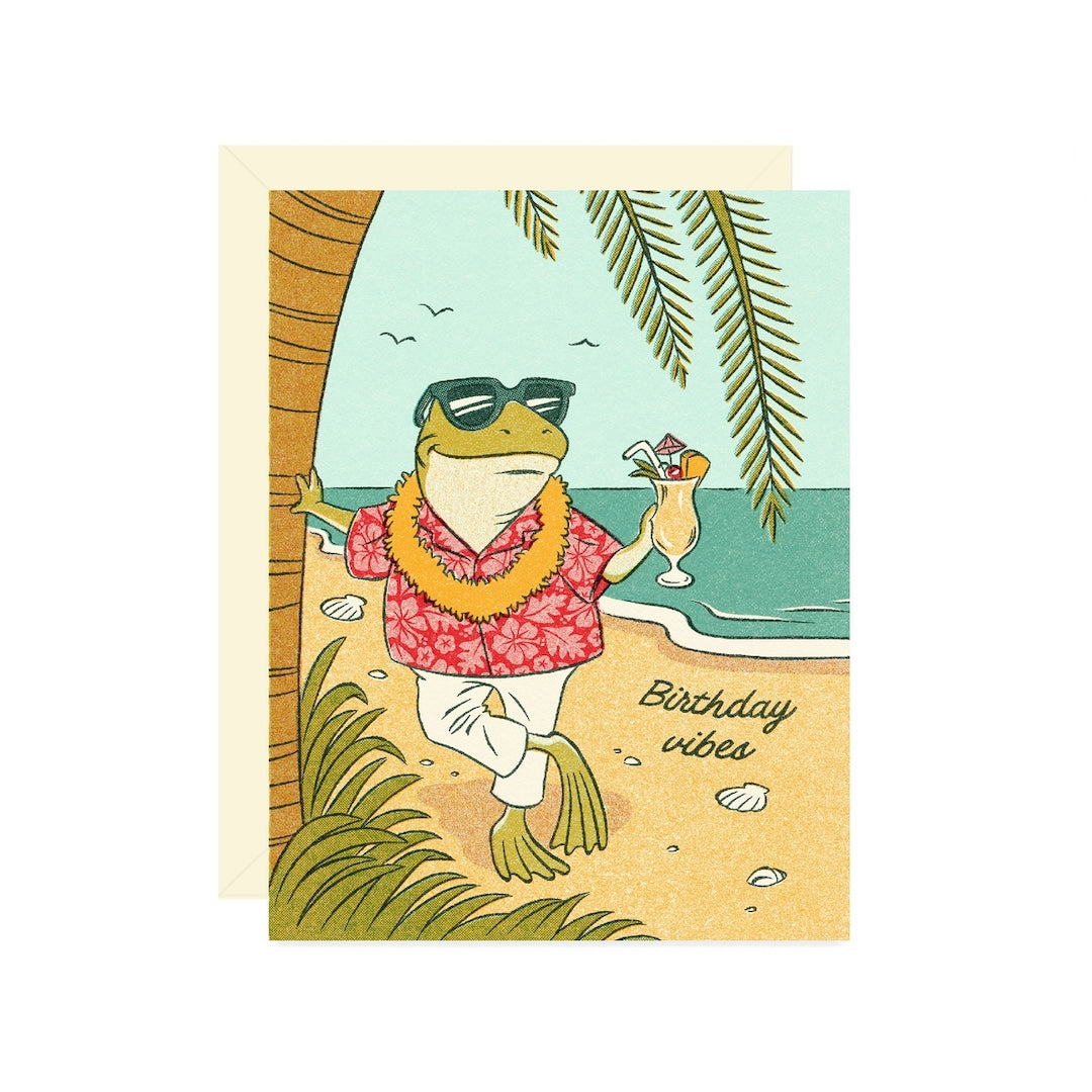 A square greeting card featuring a cartoon frog wearing sunglasses, a red Hawaiian shirt, and a yellow lei on a beach, with the text 'Birthday vibes.' A cream envelope is partially visible behind the card on a white background.