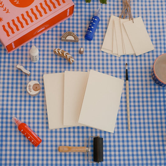 block printing items including note cards, gift tags, a paintbrush, stamps, paint, and a roller on a checkered surface