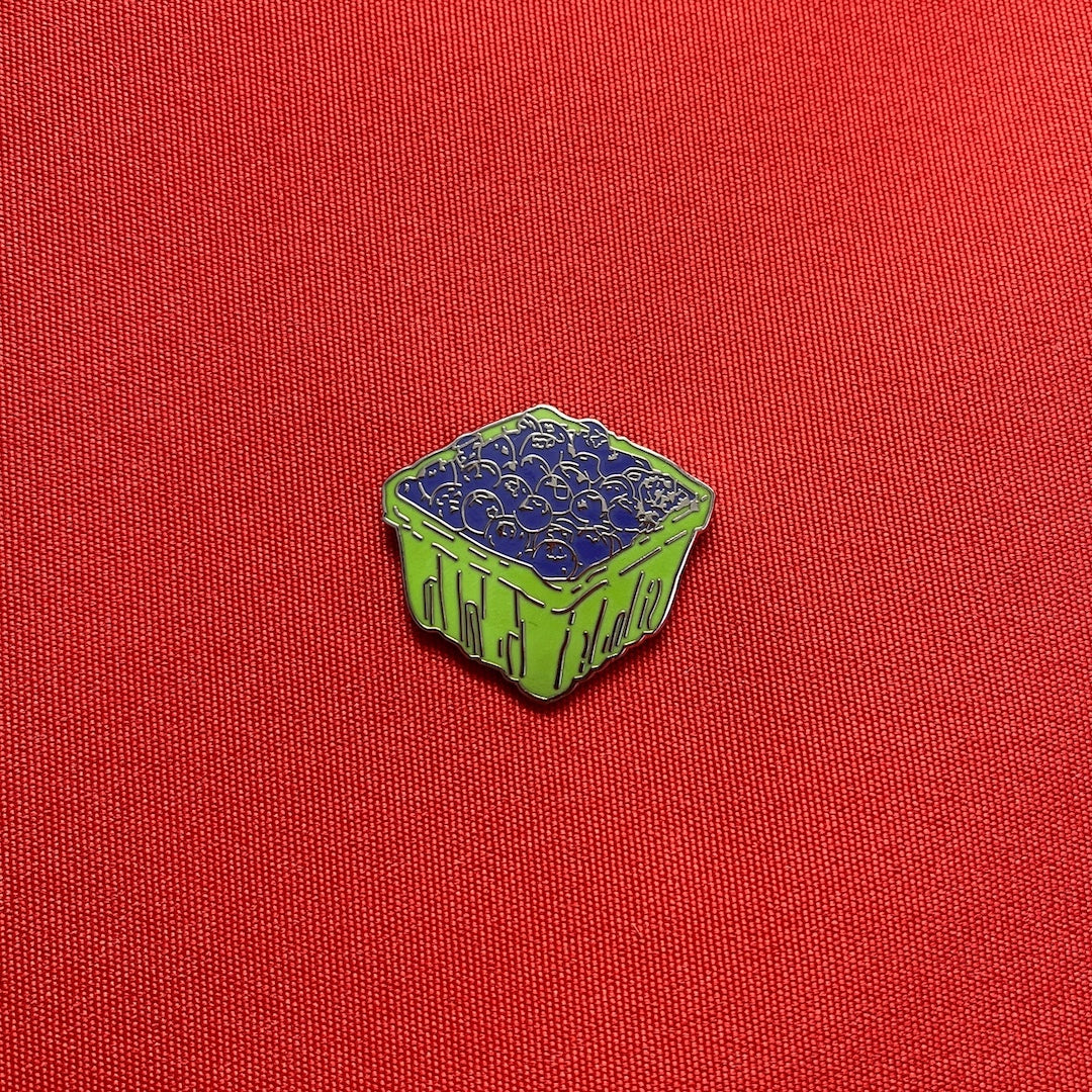 A small enamel pin shaped like a green basket filled with blueberries, placed on a textured red fabric background.