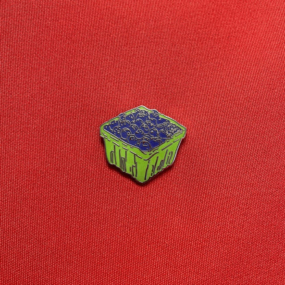 A small enamel pin shaped like a green basket filled with blueberries, placed on a textured red fabric background.