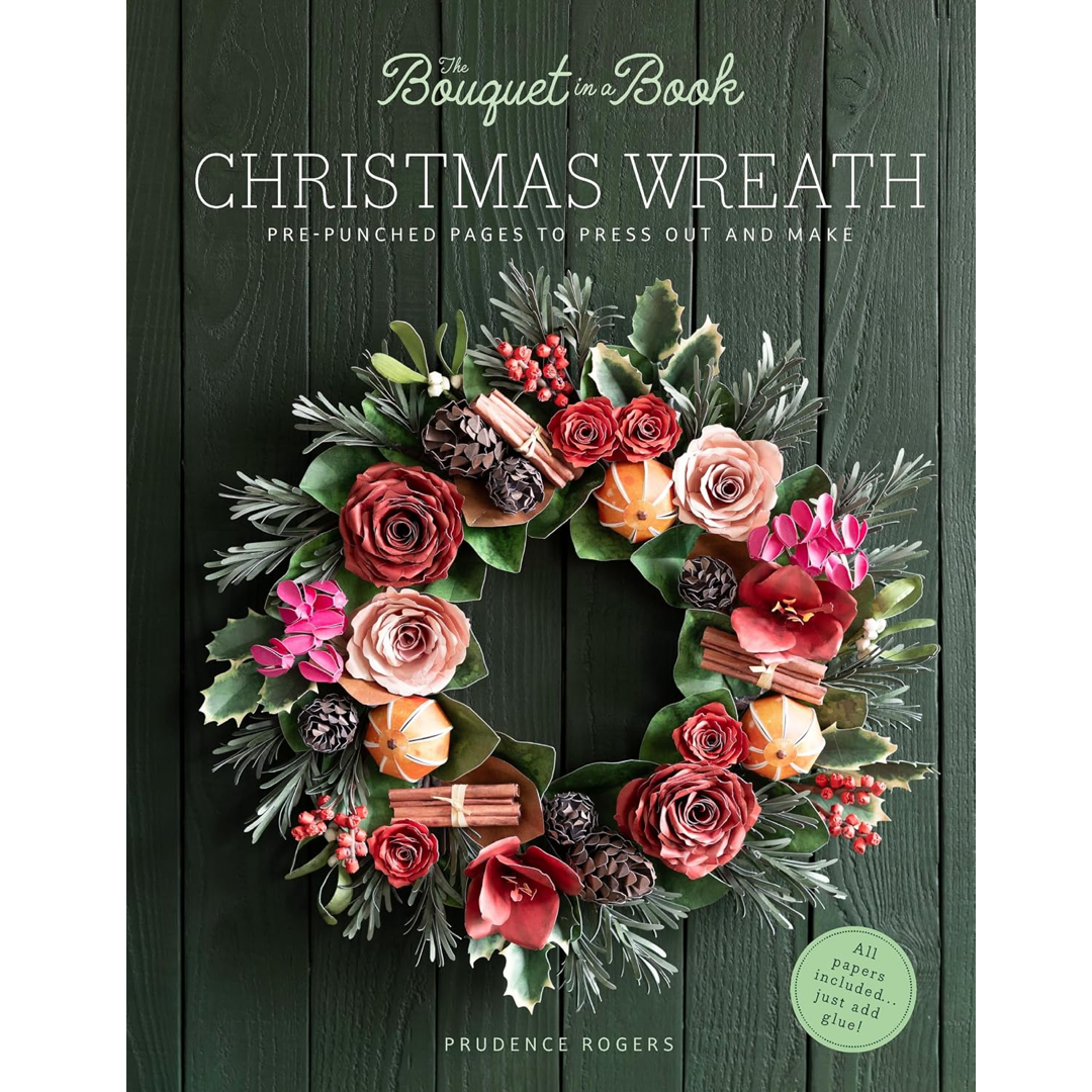 Front cover of a craft book titled "The Bouquet in a Book: Christmas Wreath" by Prudence Rogers, showing a colorful paper wreath with roses, pinecones, cinnamon sticks, berries, and greenery against a dark green wood-plank background.