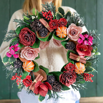 A decorative autumn wreath with artificial roses, pinecones, cinnamon sticks, berries, and mini pumpkins, held by a person wearing a cream sweater against a dark green paneled background.