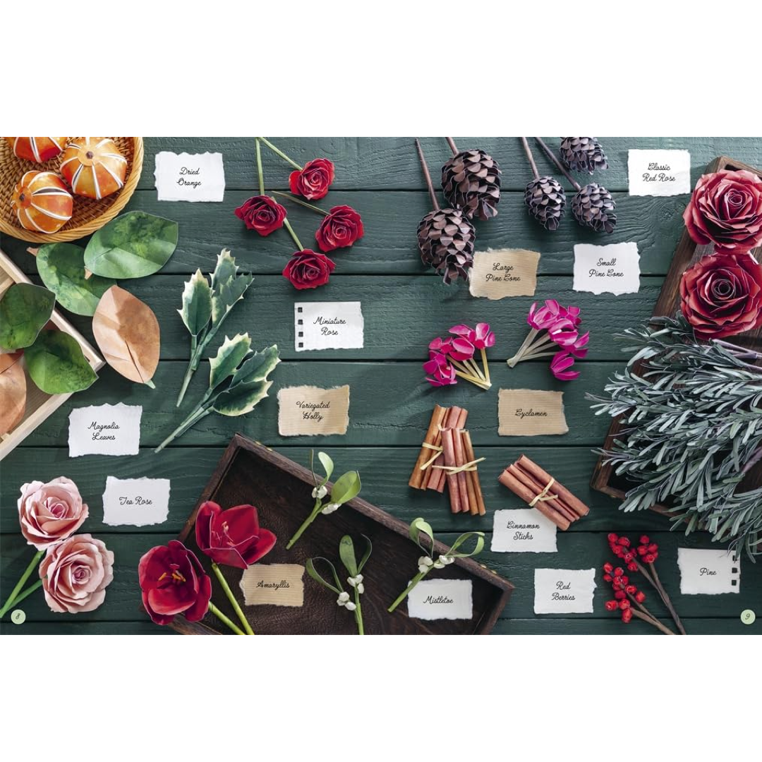 A styled flat-lay arrangement of paper floral and botanical items on a dark green wooden surface, including roses, pinecones, leaves, cinnamon sticks, berries, and small labeled cards identifying each item.