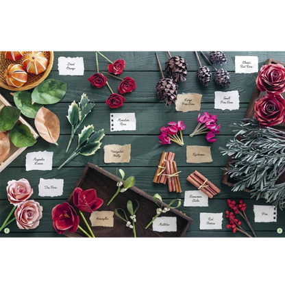 A styled flat-lay arrangement of paper floral and botanical items on a dark green wooden surface, including roses, pinecones, leaves, cinnamon sticks, berries, and small labeled cards identifying each item.