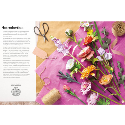 An open book spread showing a paper-flower making guide, with a bright pink sheet of paper, artificial flowers, twine, scissors, and a kraft paper background.