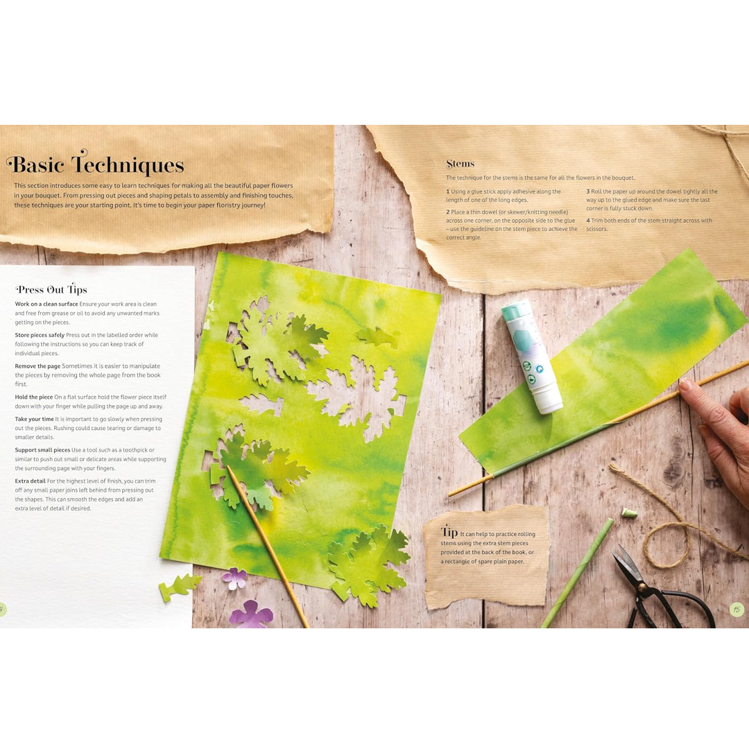 An instructional art guide page titled 'Basic Techniques' laid out on a rustic wooden table, showing examples of painted paper flowers, a glue stick, scissors, twine, and a hand holding a paintbrush.