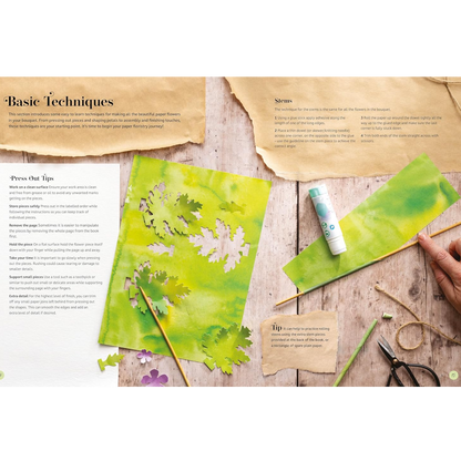 An instructional art guide page titled 'Basic Techniques' laid out on a rustic wooden table, showing examples of painted paper flowers, a glue stick, scissors, twine, and a hand holding a paintbrush.