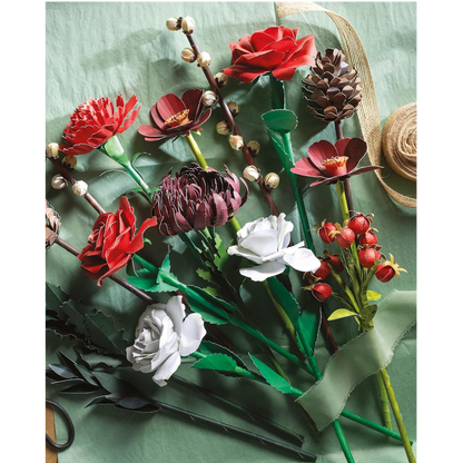A collection of artificial paper flowers and buds arranged on a textured sage green background, featuring red, white, and dark burgundy blossoms with green leaves and stems, alongside a brown twine ribbon.