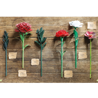 Six artificial flowers with handwritten paper labels are arranged on a rustic wooden background. The flowers include red, white, and pink blooms with green stems and leaves.