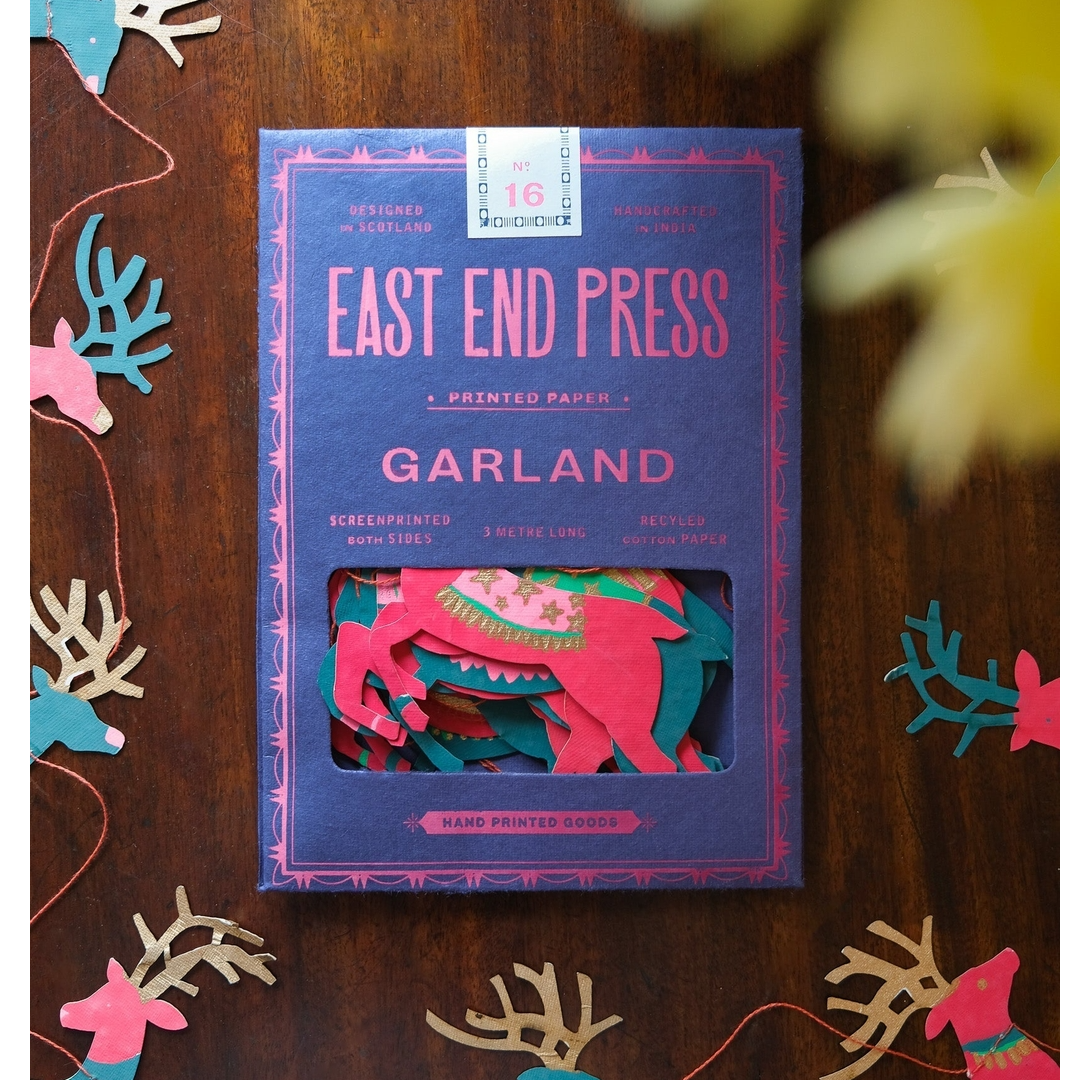 East End Press Garland printed paper packaging on a dark wood background, surrounded by colorful paper reindeer and holiday garland decorations. The box is blue with pink lettering and shows a window displaying layered pink and green paper cutouts.