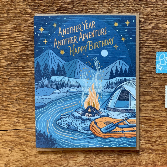 A square illustrated birthday card lying on a wooden surface, featuring a camping scene with mountains, a river, a tent, an orange inflatable raft, and the text 'ANOTHER YEAR ANOTHER ADVENTURE. Happy Birthday!'.