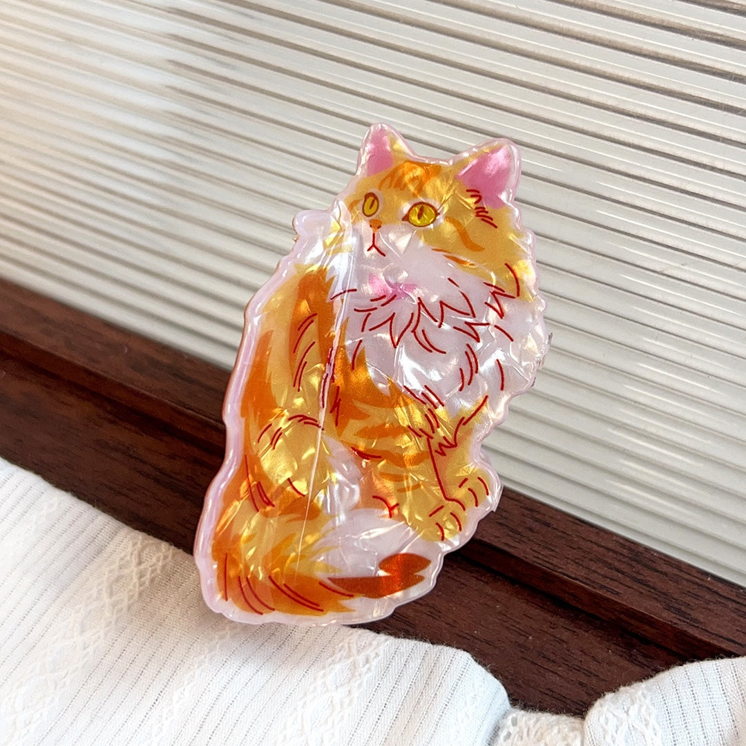 A translucent acrylic cat-shaped claw clip featuring an orange and white fluffy cat design with red line details, resting on a white textured fabric next to a dark wooden surface.