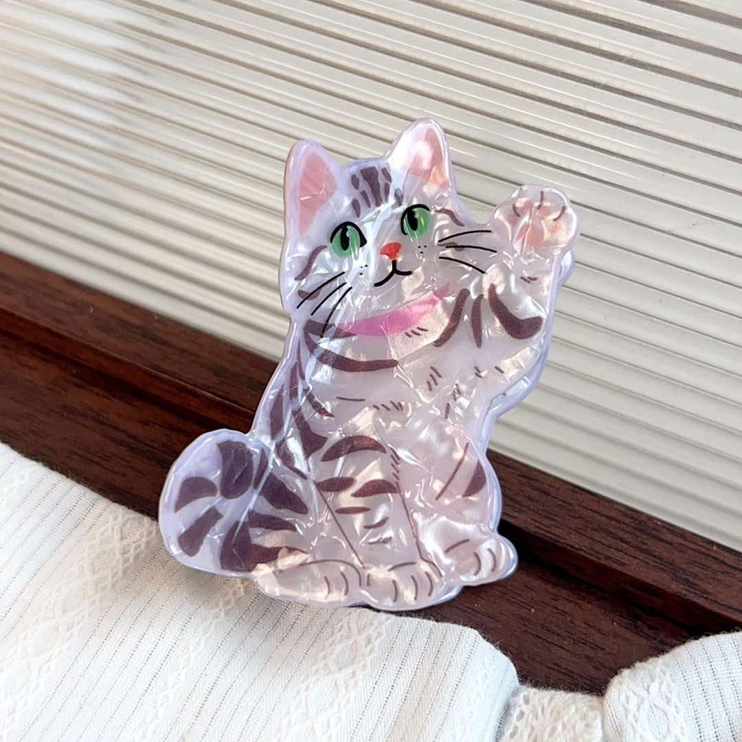 A clear acrylic cat-shaped claw clip with grey stripes,  pink collar, green eyes, and one paw is up in the air like it's waving. Shown resting on a dark wooden surface against a cream-colored slatted background.