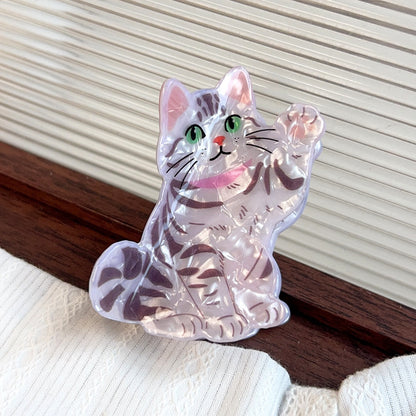 A clear acrylic cat-shaped claw clip with grey stripes,  pink collar, green eyes, and one paw is up in the air like it's waving. Shown resting on a dark wooden surface against a cream-colored slatted background.