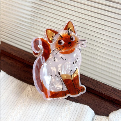 A clear acrylic cat-shaped claw clip with an orange and white cat illustration, resting on a white textured fabric next to a dark wooden surface.