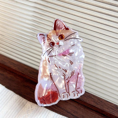 A translucent acrylic claw clip shaped like a sitting cat, featuring pink, red, and orange details with dark outlines, resting on a dark wooden surface against a cream-colored slatted background.