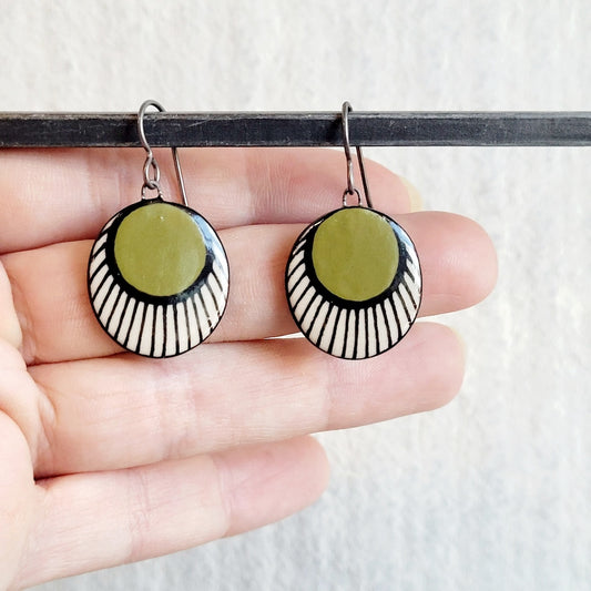 Round earrings with green, white, and black design held on a hand against a neutral background