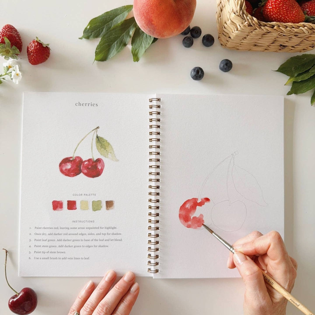 Open watercolor workbook with cherry illustration, hand holding a paintbrush, and fruits on a white surface