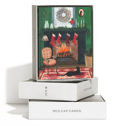 A boxed card set displayed on a white background. The top box shows an illustrated Christmas fireplace scene with stockings, a fire, rug, chair, wreath, and candlesticks.